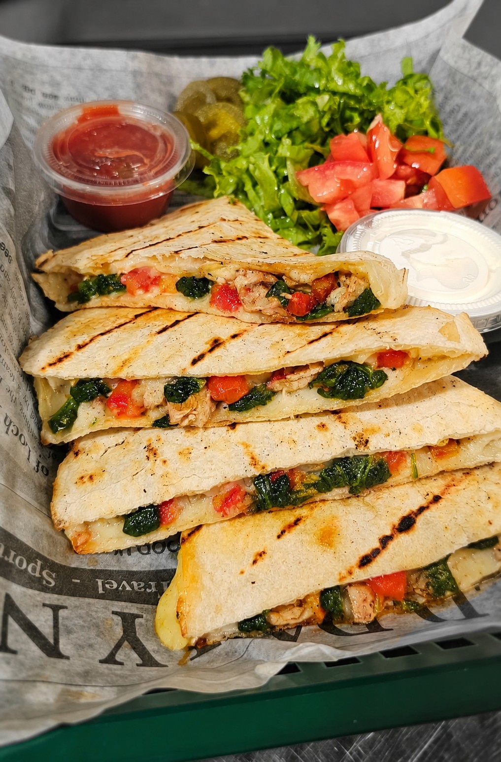 Three grilled quesadilla wedges filled with vegetables and chicken, served with salsa, sour cream, lettuce, diced tomato, and jalapeños on a tray lined with printed paper.