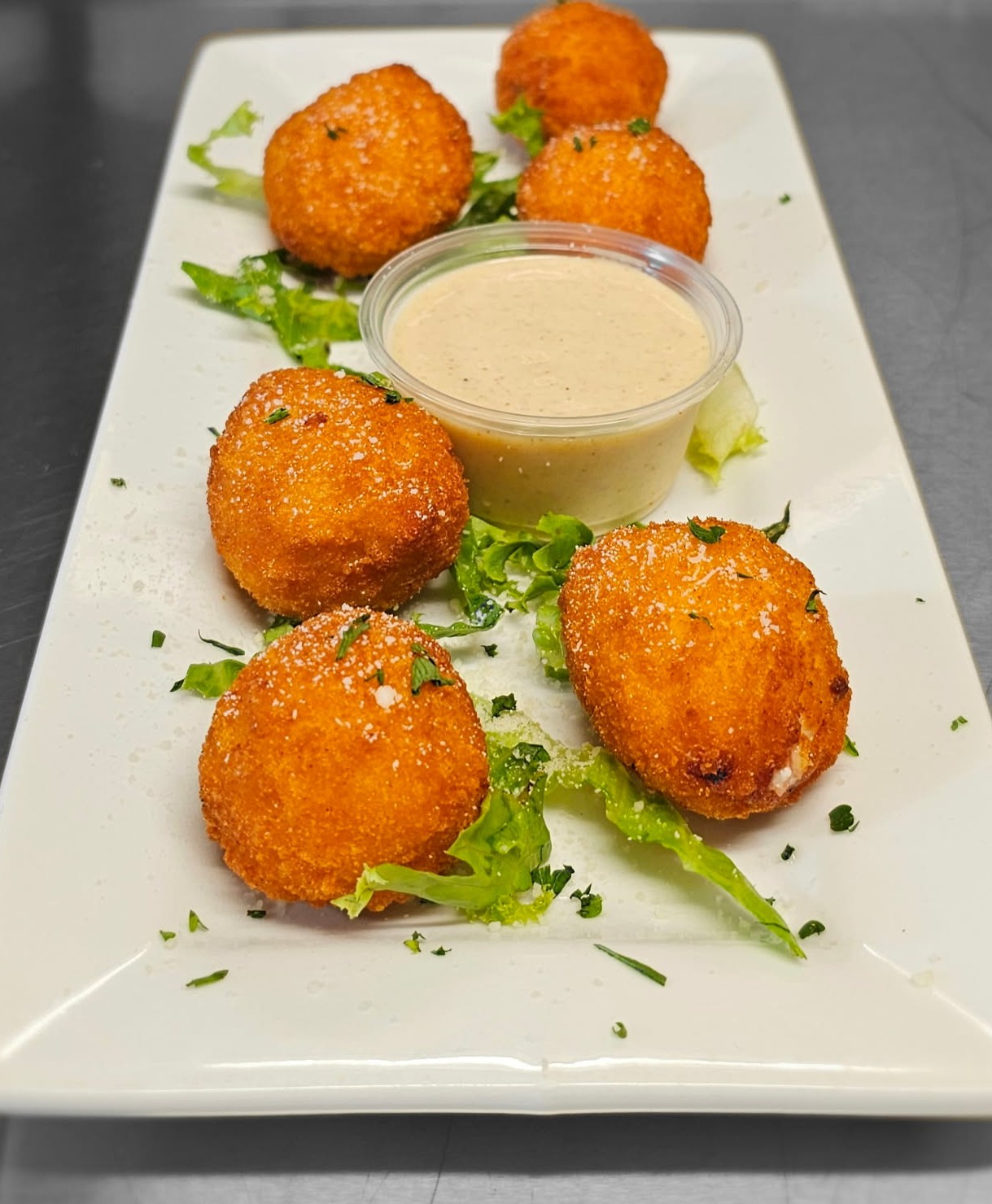 Six golden-brown fried balls are arranged on a white rectangular plate with greens and a small container of creamy dipping sauce in the center.