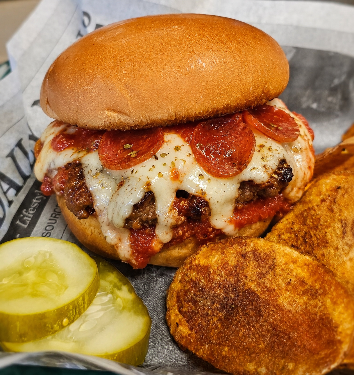 A cheeseburger topped with pepperoni and melted cheese, served with potato chips and pickle slices on a sheet of paper.